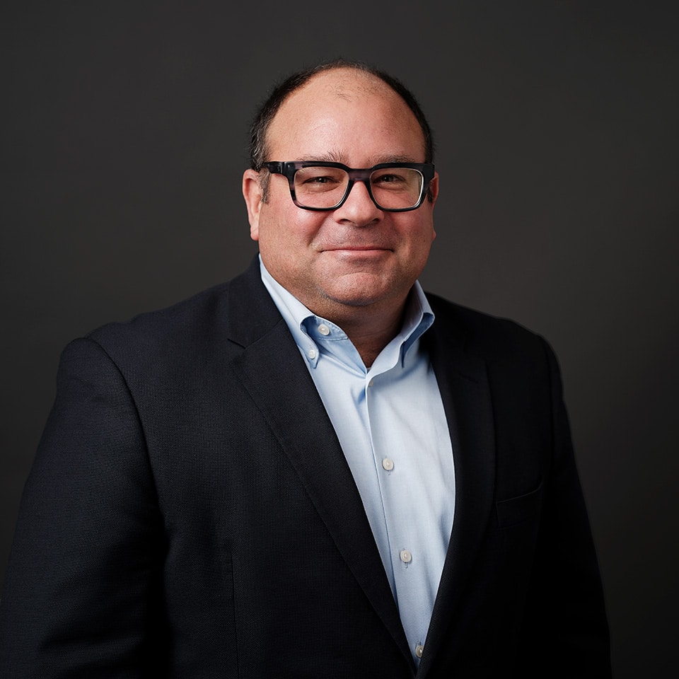 Headshot of John Peluso in a navy blazer with a dark background.