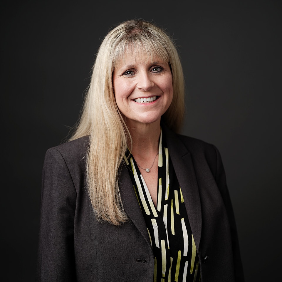 Headshot of Karen Coker with blonde hair and wearing a gray blazer in front of a dark background.